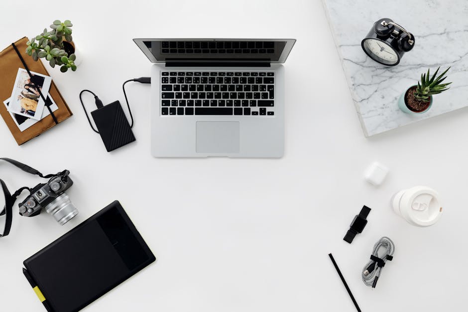 Top view of a minimalist workspace featuring a laptop, camera, and various electronic accessories on a white table.