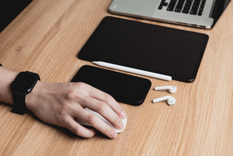 A hand using a mouse with a laptop, tablet, smartphone, and earphones on a wooden desk.